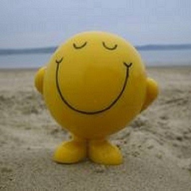 A yellow round smiley face figure with closed eyes and a big smile standing on sandy beach with a calm sea and cloudy sky in the background.
