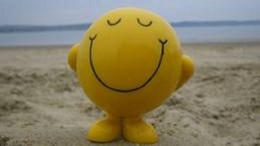 A yellow round smiley face figure with closed eyes and a big smile standing on sandy beach with a calm sea and cloudy sky in the background.