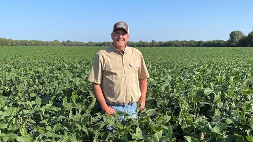 A man wearing a beige shirt, blue jeans, and a gray cap stands smiling in the middle of a lush green soybean field under a clear blue sky with trees in the distant background.