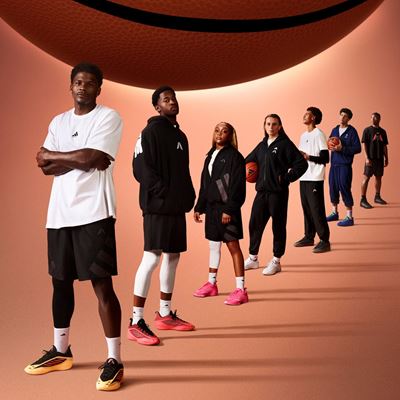A diverse group of seven young basketball players standing in a diagonal line on a brown background with a large basketball hovering above them. They are dressed in sporty basketball attire, including shorts, hoodies, and sneakers, with some holding basketballs. The lighting creates long shadows behind them, emphasizing their confident and focused poses.