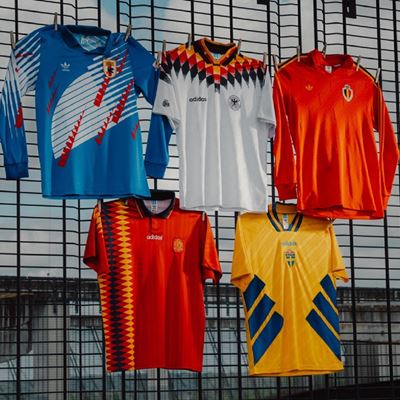 Five vintage soccer jerseys hanging on a metal fence outdoors, featuring various national team designs and colors including blue, white, red, orange, and yellow with distinct patterns and Adidas branding.