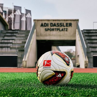 A close-up of a Bundesliga soccer ball resting on green grass in front of the entrance tunnel of a stadium named "Adi Dassler Sportplatz," with empty concrete bleachers and Adidas banners visible in the background.