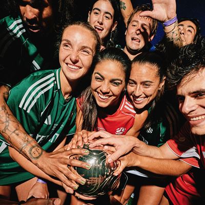 A diverse group of smiling young athletes wearing green and red sports jerseys gather closely together, joyfully holding a shiny metallic ball in the center, celebrating teamwork and camaraderie.