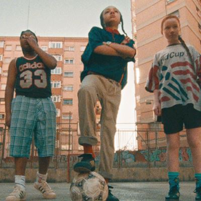 Three children standing on an outdoor basketball court in an urban setting with tall apartment buildings in the background. The child on the left is wearing a black Adidas sleeveless shirt with the number 33 and plaid shorts, covering their mouth with one hand. The child in the middle is standing confidently with arms crossed, one foot resting on a soccer ball, wearing a dark shirt and beige pants. The child on the right is wearing a loose sports jersey and black shorts, standing with hands by their sides. The scene has a warm, nostalgic tone.