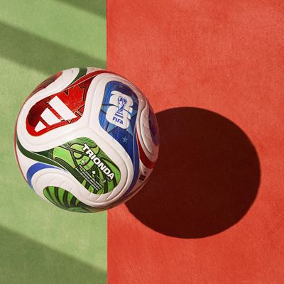 A colorful Adidas Trionda FIFA soccer ball casting a shadow on a surface divided into green and red sections, with sunlight creating diagonal shadows on the green side.