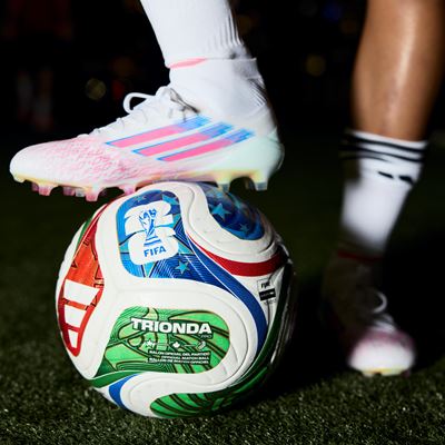 Close-up of a soccer player's legs wearing white cleats with pink and blue accents and white socks with black stripes, standing on a green artificial turf field with one foot resting on a colorful FIFA-approved soccer ball.