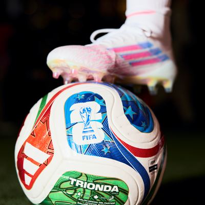 A close-up image of a colorful FIFA Trionda soccer ball on a grass field with a player's foot wearing a white cleat and white sock resting on top of the ball.