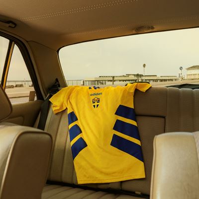 A yellow Adidas sports jersey with blue diagonal stripes is draped over the back seat of a beige car interior, with a view of a pier and ocean visible through the rear window.