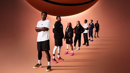 A diverse group of seven young basketball players standing in a diagonal line on a brown background with a large basketball hovering above them. They are dressed in sporty basketball attire, including shorts, hoodies, and sneakers, with some holding basketballs. The lighting creates long shadows behind them, emphasizing their confident and focused poses.