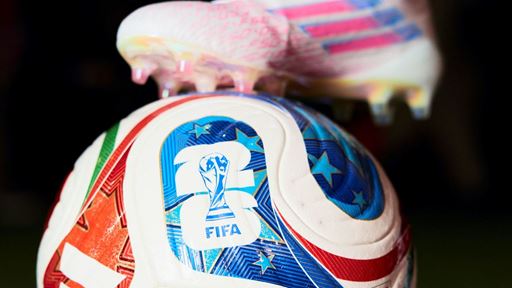 Close-up of a colorful FIFA Trionda soccer ball on a grass field with a player's foot wearing white cleats and white socks resting on top of the ball.