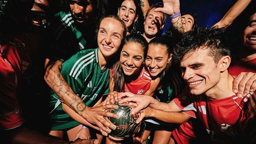 A diverse group of smiling young athletes wearing green and red sports jerseys gather closely together, joyfully holding a shiny metallic ball in the center, celebrating teamwork and camaraderie.