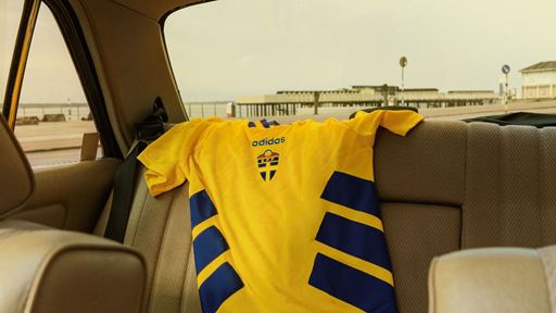 A yellow Adidas sports jersey with blue diagonal stripes is draped over the back seat of a beige car interior, with a view of a pier and cloudy sky visible through the rear window.