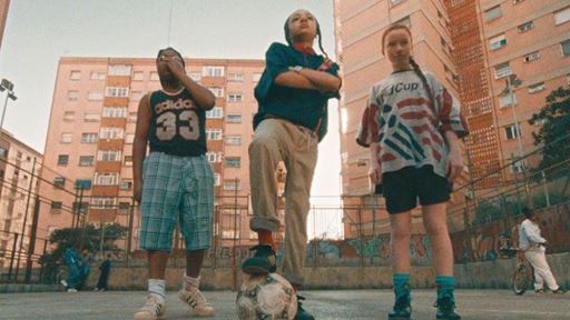 Three children standing on an outdoor basketball court in an urban setting with tall apartment buildings in the background. The child on the left is wearing a black Adidas sleeveless shirt with the number 33 and plaid shorts, covering their mouth with one hand. The child in the middle is standing confidently with arms crossed, one foot resting on a soccer ball, wearing a dark shirt and beige pants. The child on the right is wearing a loose sports jersey and black shorts, standing with hands by their sides. The scene has a warm, nostalgic tone.