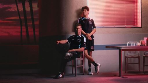 Two men wearing black sportswear with Audi and Revolut logos pose in a dimly lit room with red lighting. One man is sitting on a chair with one leg crossed over the other, while the other man stands behind him with his hands clasped. The room features a table with chairs and a window with closed blinds, casting a red glow. The background includes a dark wall and a sunset scene with palm trees.