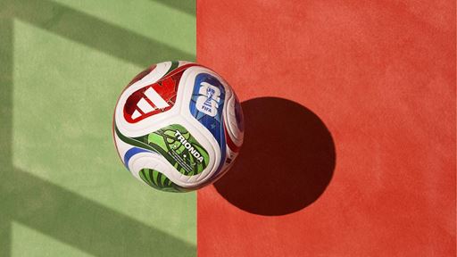 A colorful Adidas Trionda FIFA soccer ball casting a shadow on a surface divided into green and red sections, with sunlight creating diagonal shadows on the green side.