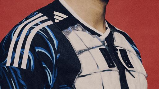 Close-up of a person wearing a black and blue Adidas sports jersey with white stripes on the shoulder and a transparent chest protector against a red background.