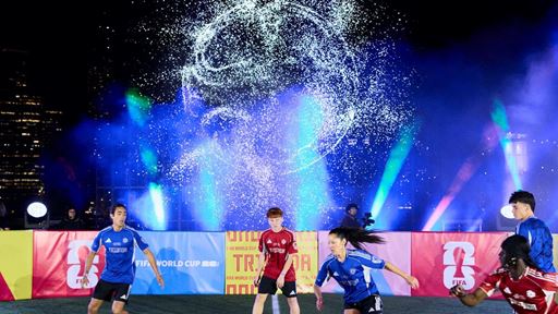 A group of young soccer players actively playing a match on a well-lit field at night, with vibrant blue and purple stage lights and a dramatic water splash effect in the background, creating an energetic and dynamic atmosphere.