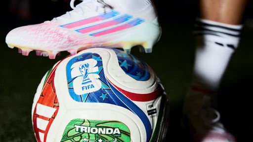 Close-up of a soccer player's legs wearing white cleats with pink and blue accents and white socks with black stripes, standing on a green artificial turf field with one foot resting on a colorful FIFA-approved soccer ball.