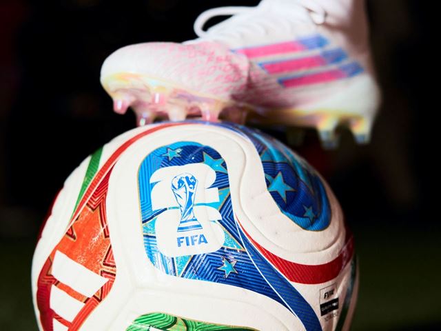 Close-up of a colorful FIFA Trionda soccer ball on a grass field with a player's foot wearing white cleats and white socks resting on top of the ball.