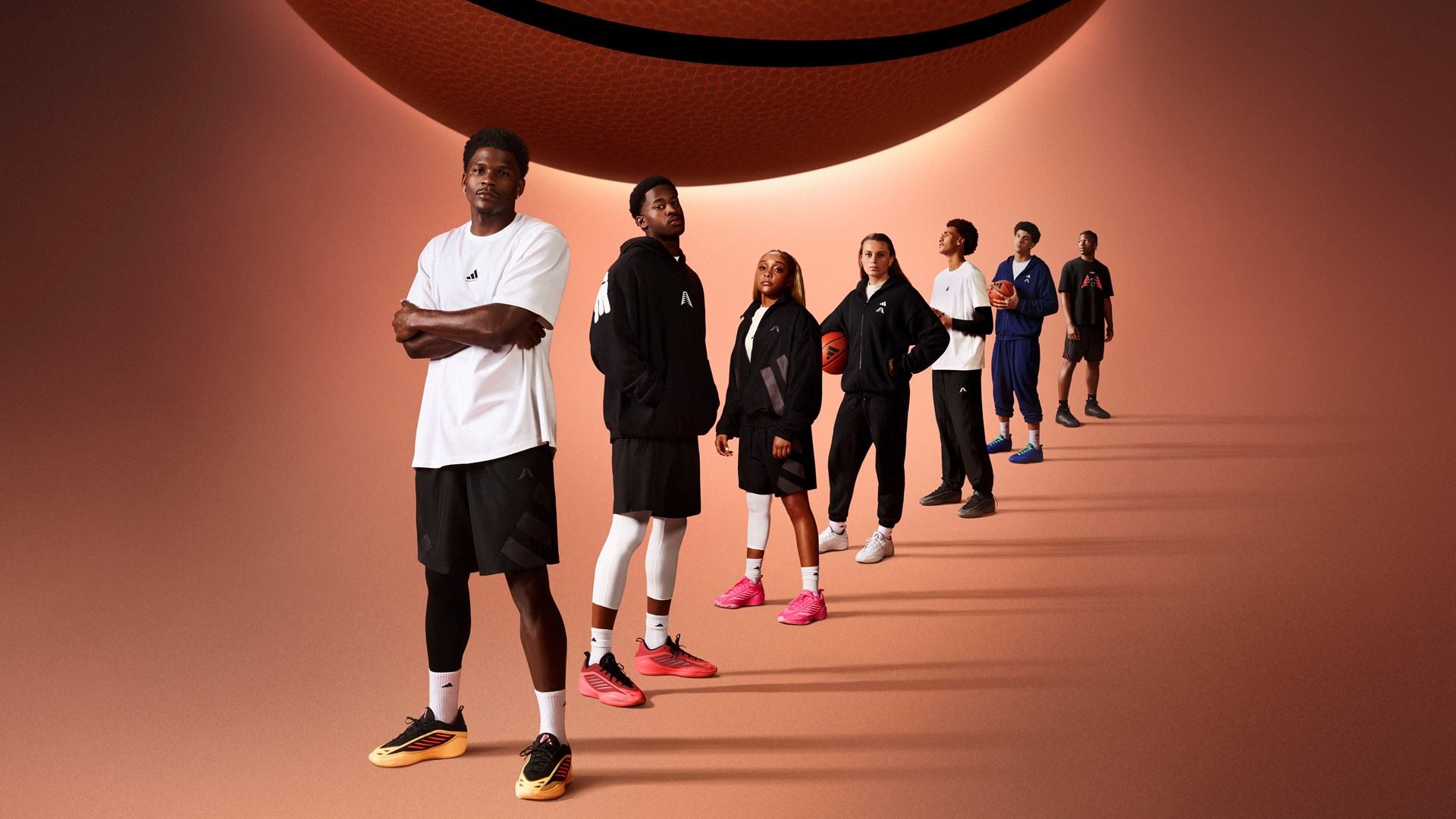 A diverse group of seven young basketball players standing in a diagonal line on a brown background with a large basketball hovering above them. They are dressed in sporty basketball attire, including shorts, hoodies, and sneakers, with some holding basketballs. The lighting creates long shadows behind them, emphasizing their confident and focused poses.